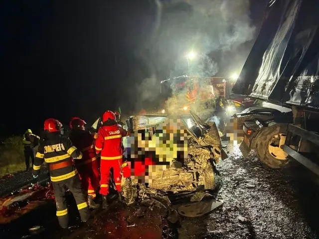 Pompieri în uniforme reflectorizante lucrează lângă un autoturism distrus și incendiat lângă un camion mare pe un drum noaptea.
