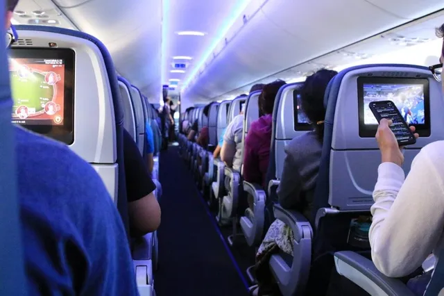 Pasageri stau așezați în scaunele unui avion, utilizând ecrane individuale de divertisment montate pe spătarele scaunelor din fața lor.