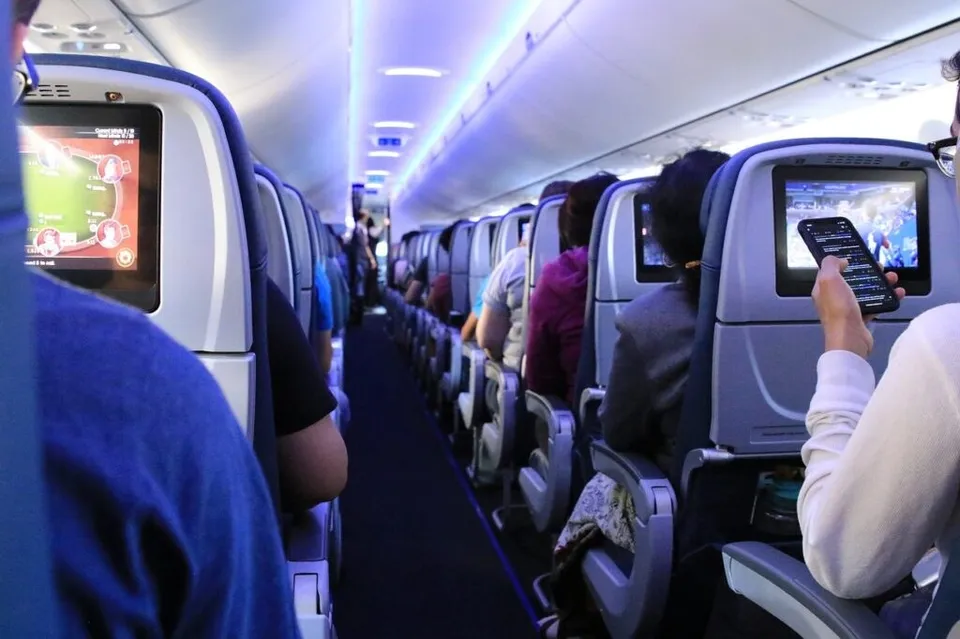 Pasageri stau așezați în scaunele unui avion, utilizând ecrane individuale de divertisment montate pe spătarele scaunelor din fața lor.