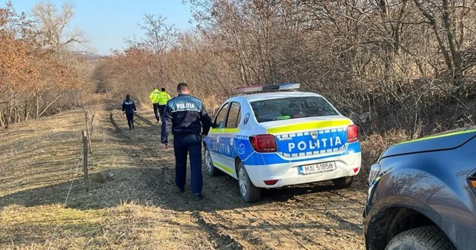 O mașină de poliție albastră și albă stă pe un drum de pământ lângă trei persoane purtând haine colorate într-o zonă cu vegetație uscată.