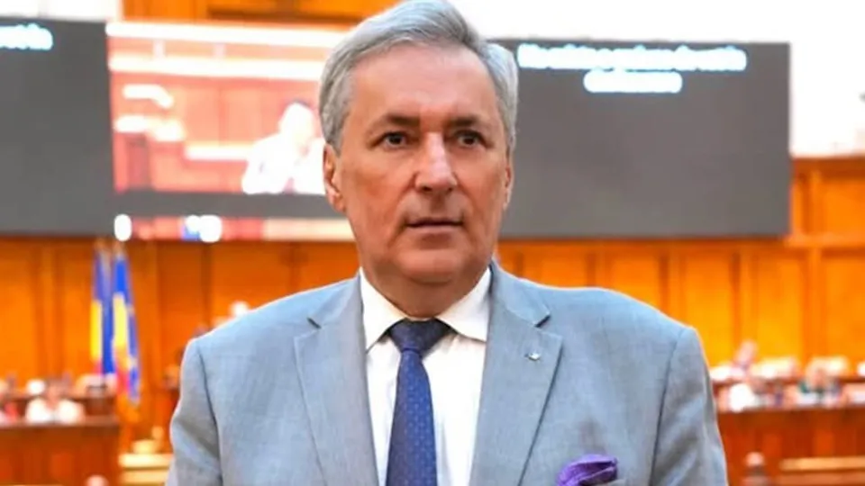 Un bărbat cu părul cărunt, purtând sacou gri, cămașă albă și cravată albastră, stă în fața unui ecran mare într-o sală cu panouri de lemn.