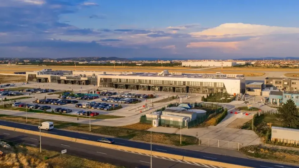 O vedere aeriană a unui terminal aeroportuar cu o parcare plină de mașini și o autostradă în prim-plan sub un cer cu nori.