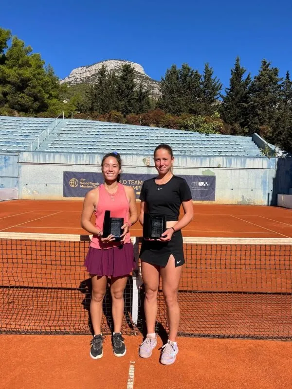 Două femei stau pe un teren de tenis de culoare portocalie, purtând haine sportive și ținând plăcuțe de premiu negre.