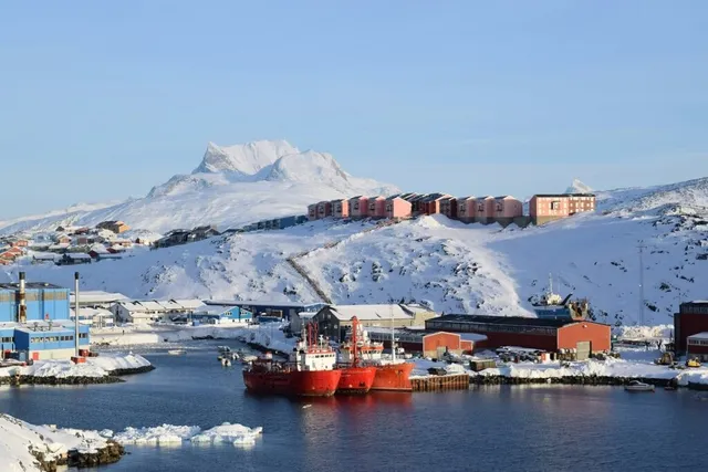Un port înghețat cu nave roșii, clădiri colorate și un munte înzăpezit în fundal.