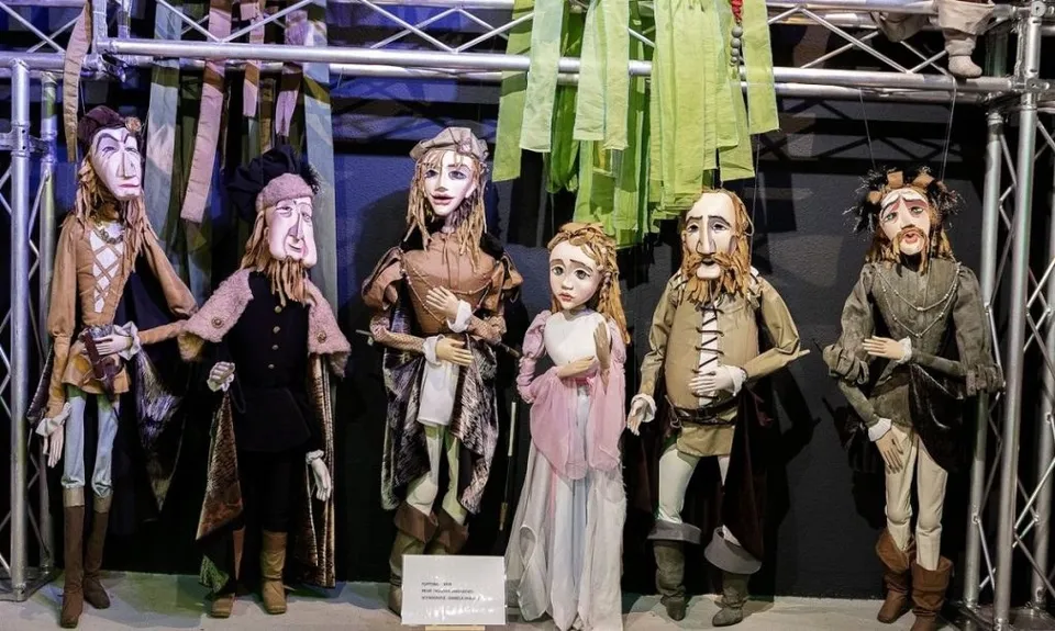 Șase marionete de dimensiuni medii, îmbrăcate în haine colorate, sunt suspendate pe un cadru metalic în fața unui fundal textil albastru și verde.