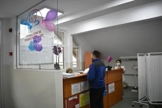 Un bărbat cu o jachetă albastră stă în fața unui birou de recepție într-o încăpere cu baloane colorate și un panou informativ.
