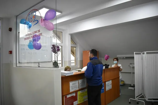 Un bărbat cu o jachetă albastră stă în fața unui birou de recepție într-o încăpere cu baloane colorate și un panou informativ.