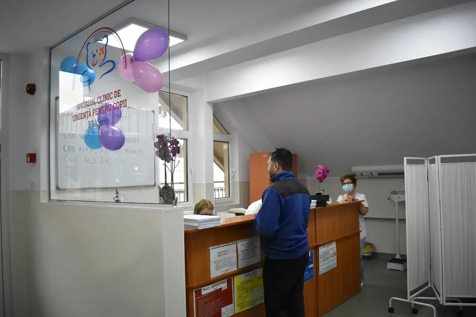 Un bărbat cu o jachetă albastră stă în fața unui birou de recepție într-o încăpere cu baloane colorate și un panou informativ.