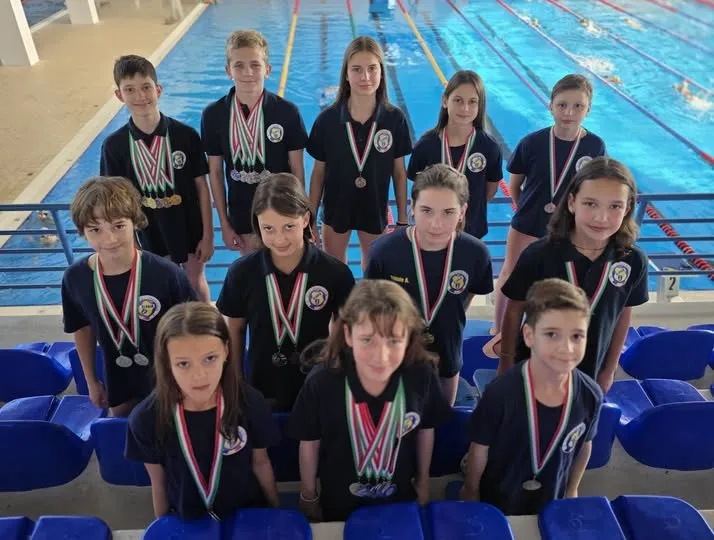 Un grup de copii îmbrăcați în echipamente sportive negre cu detalii verzi stau în fața unei piscine, purtând medalii în jurul gâtului.