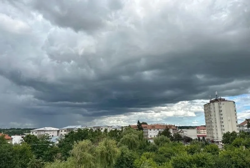 Un cer acoperit de nori întunecați se întinde deasupra unor clădiri albe și a unui copăret verde dens.
