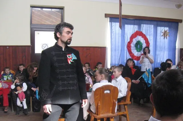 Un bărbat cu barbă, îmbrăcat într-o haină tradițională neagră, stă în fața unui grup de copii și adulți, într-o sală decorată cu un cocardă tricoloră.