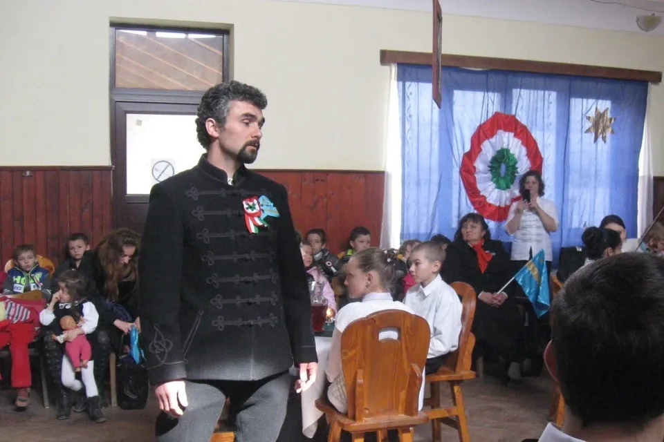 Un bărbat cu barbă, îmbrăcat într-o haină tradițională neagră, stă în fața unui grup de copii și adulți, într-o sală decorată cu un cocardă tricoloră.
