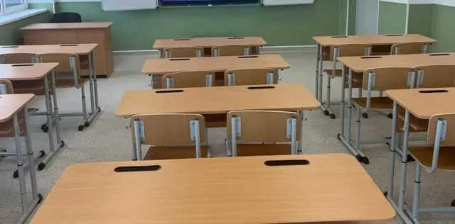 O sală de clasă cu mai multe birouri din lemn și scaune metalice dispuse pe rânduri pe un podea gri.