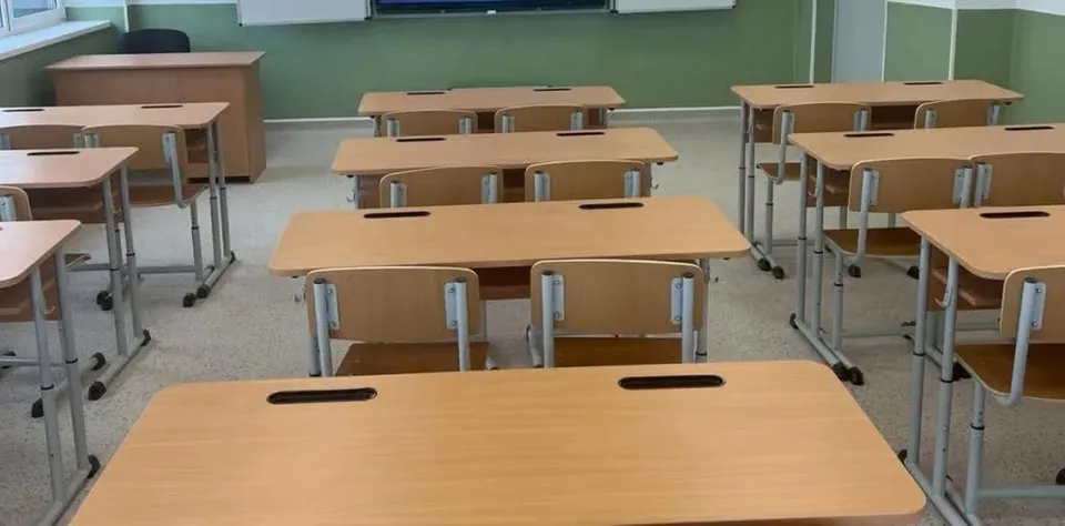 O sală de clasă cu mai multe birouri din lemn și scaune metalice dispuse pe rânduri pe un podea gri.