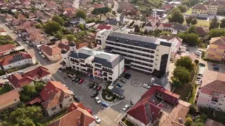 O vedere aeriană prezintă un complex de clădiri cu acoperișuri în tonuri de gri și gri-albastru, înconjurat de case cu acoperișuri roșii și vegetație.