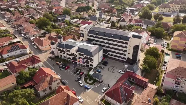 O vedere aeriană prezintă un complex de clădiri cu acoperișuri în tonuri de gri și gri-albastru, înconjurat de case cu acoperișuri roșii și vegetație.