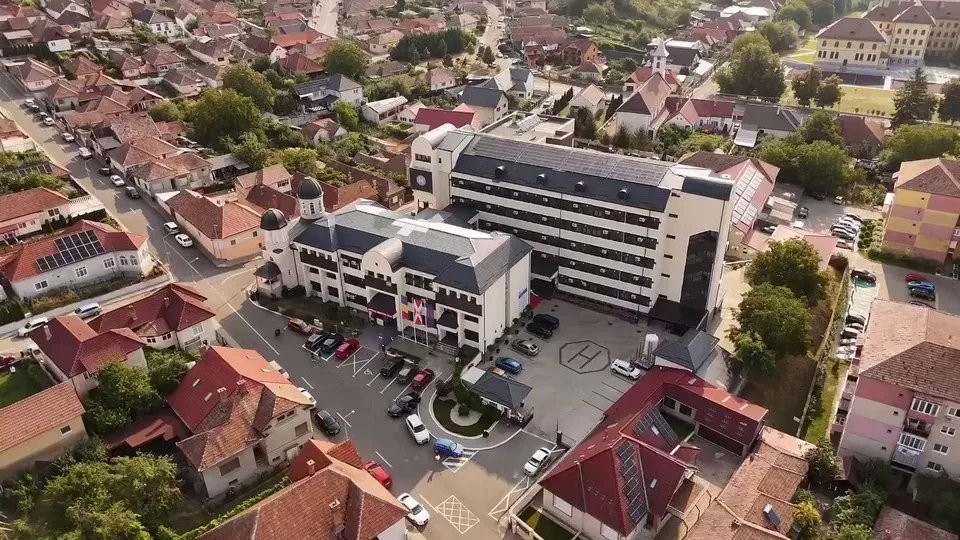 O vedere aeriană prezintă un complex de clădiri cu acoperișuri în tonuri de gri și gri-albastru, înconjurat de case cu acoperișuri roșii și vegetație.