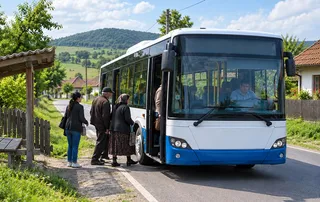 Oameni stau pe marginea unui drum lângă un autobuz albastru și alb oprit, într-un peisaj rural cu dealuri verzi sub un cer înnorat.