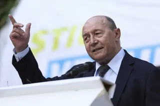 Un bărbat cu părul rarefacește, purtând sacou închis și cămașă albastră, vorbește la un microfon ridicând mâna dreaptă cu degetul arătător în sus.