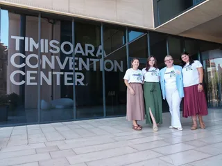 Patru femei stau în rând în fața unui clădire cu inscripția Timisoara Convention Center, purtând tricouri albe și fuste sau pantaloni lungi.