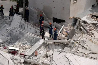 Mai mulți salvatori în uniforme de lucru și casci colorați se află printre dărâmăturile unei clădiri prăbușite.