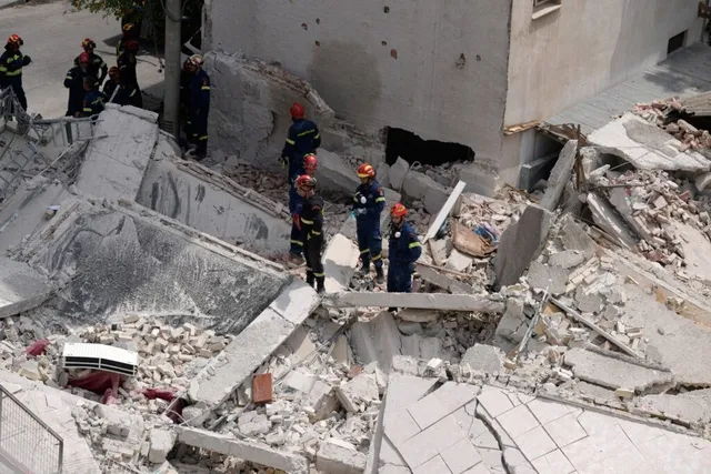 Mai mulți salvatori în uniforme de lucru și casci colorați se află printre dărâmăturile unei clădiri prăbușite.