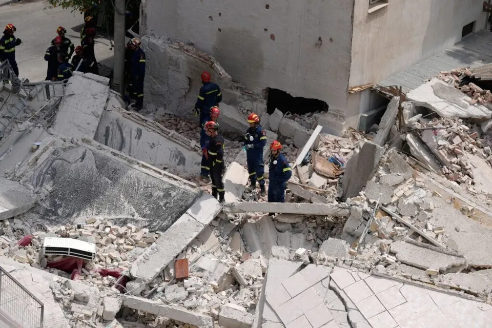 Mai mulți salvatori în uniforme de lucru și casci colorați se află printre dărâmăturile unei clădiri prăbușite.