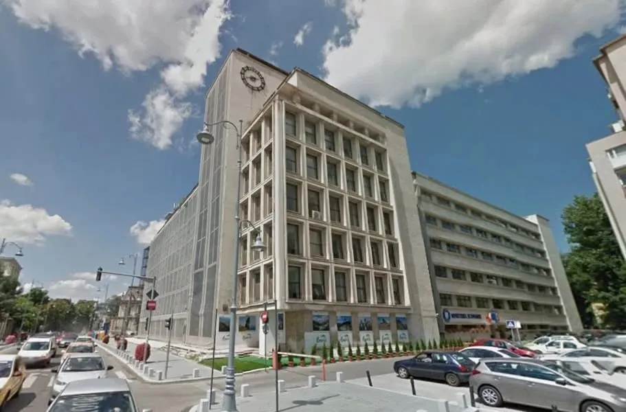 O clădire înaltă de beton cu multe ferestre este situată lângă un drum pe care circulă diverse mașini sub un cer cu nori.