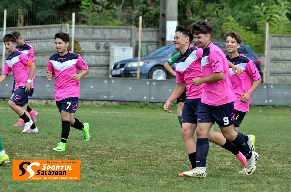 Cinci tineri jucători de fotbal în echipamente roz și negre aleargă pe un teren de iarbă verde.