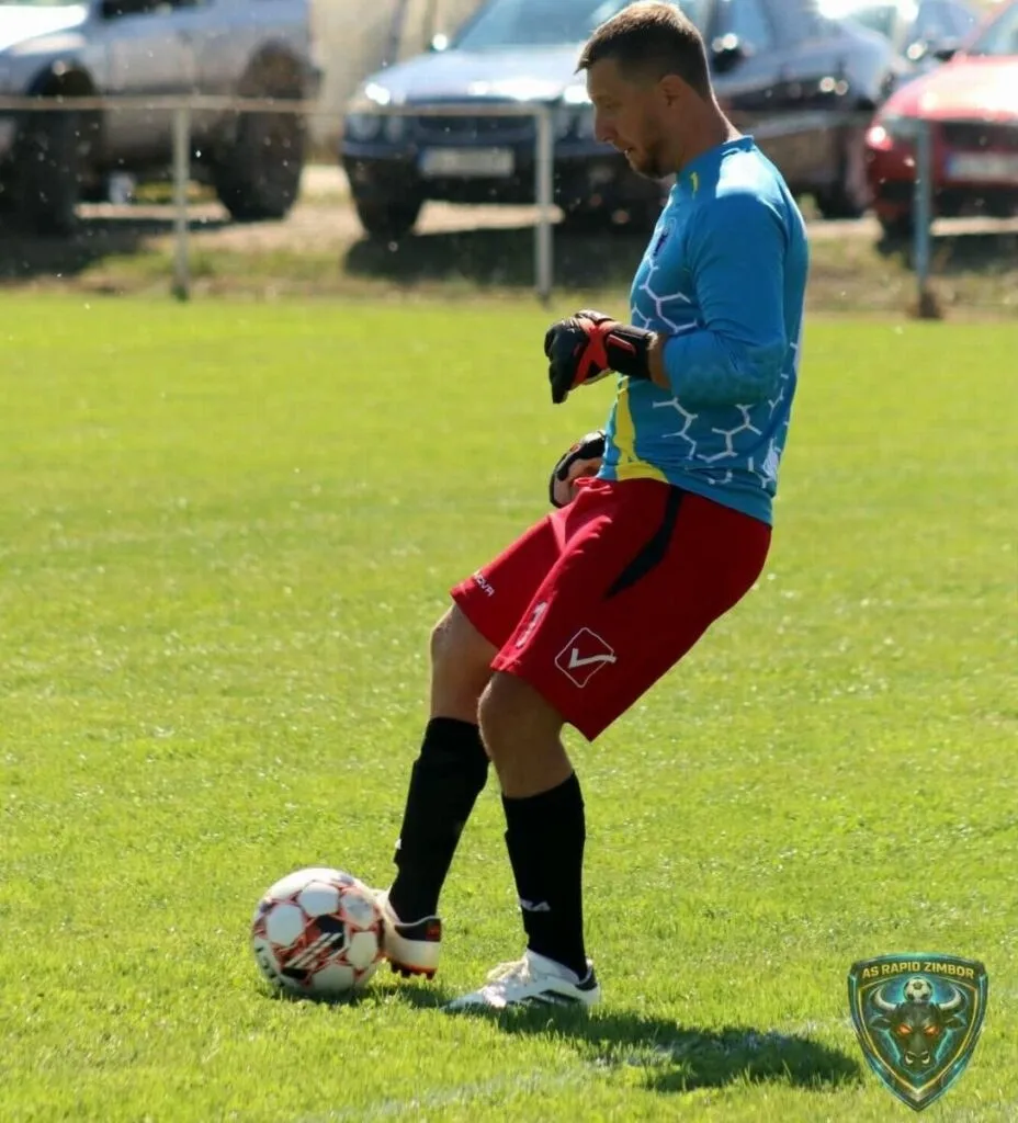 Un fotbalist cu mânuși de portar, tricou albastru și pantaloni roșii, stă pe terenul de fotbal lângă un balon.