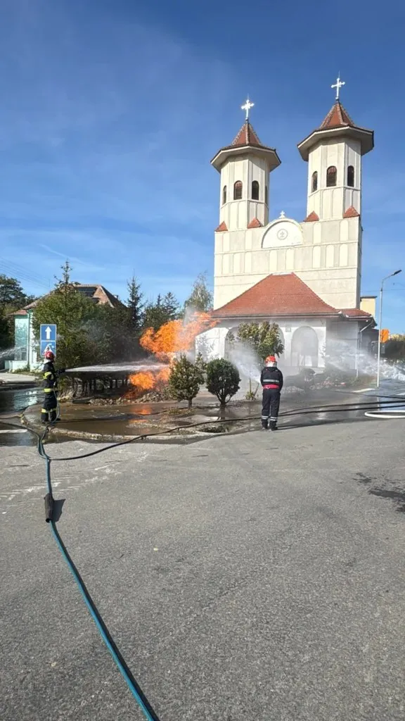 Pompieri în echipament de protecție stau pe un drum lângă flăcări și fum care ies de sub o structură metalică lângă o biserică.