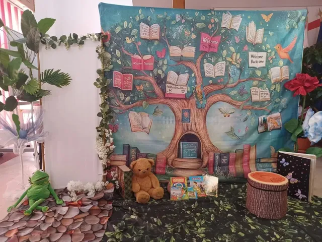 Un decor cu un copac pictat pe fundal albastru, având cărți deschise pe ramuri, alături de un urs de pluș și o plantă verde.