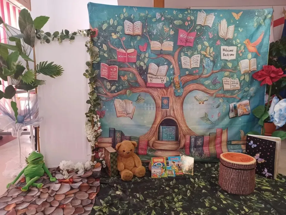Un decor cu un copac pictat pe fundal albastru, având cărți deschise pe ramuri, alături de un urs de pluș și o plantă verde.