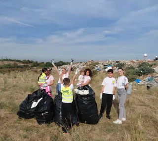 Un grup de persoane stă pe un câmp de iarbă uscată lângă saci negri de gunoi, având un morman de deșeuri în fundal.
