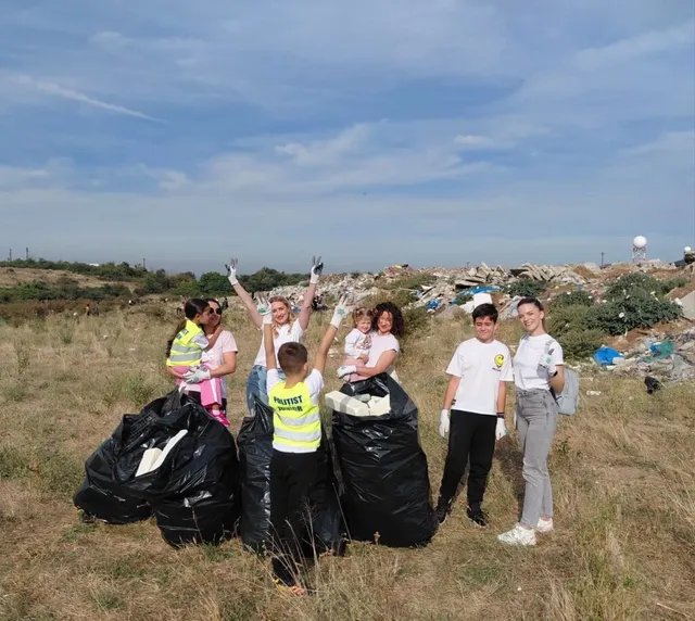 Un grup de persoane stă pe un câmp de iarbă uscată lângă saci negri de gunoi, având un morman de deșeuri în fundal.