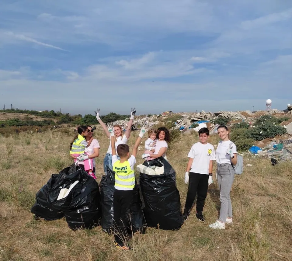 Un grup de persoane stă pe un câmp de iarbă uscată lângă saci negri de gunoi, având un morman de deșeuri în fundal.