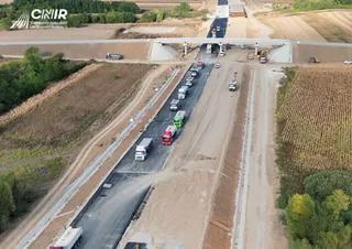 O vedere aeriană prezintă o porțiune de autostradă în construcție cu mașini, camioane și utilaje de lucru pe suprafețe asfaltate și de pământ.