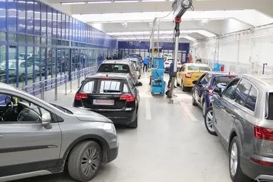 Mai multe mașini sunt parcate într-un spațiu interior cu podea albă, lângă un perete de sticlă și echipamente tehnice suspendate.