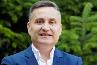 Un bărbat cu părul scurt și cămașă albă sub un sacou albastru zâmbește în fața unor frunze verzi.