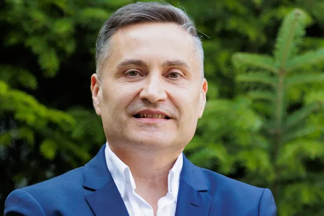 Un bărbat cu părul scurt și cămașă albă sub un sacou albastru zâmbește în fața unor frunze verzi.