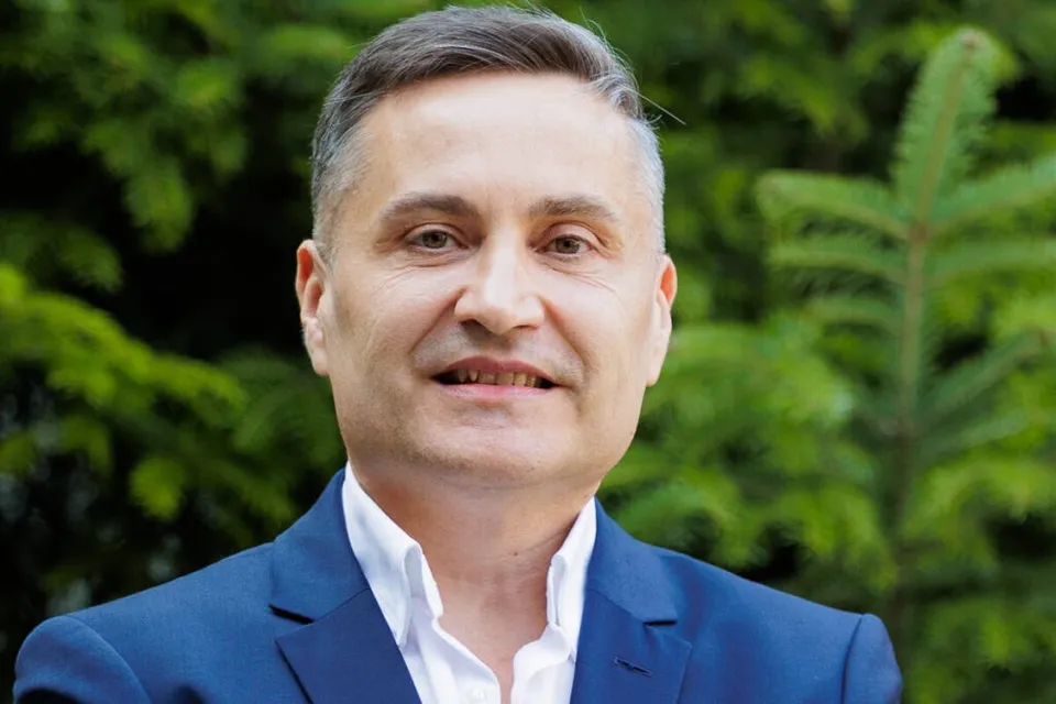 Un bărbat cu părul scurt și cămașă albă sub un sacou albastru zâmbește în fața unor frunze verzi.