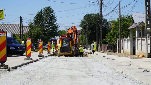 Un utilaj de construcții portocaliu și mai mulți lucrători cu veste reflectorizante se află pe un drum de pietriș lângă o clădire.