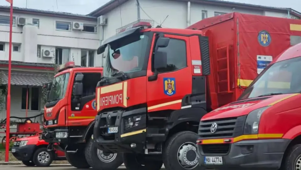 Trei vehicule roșii de intervenție, inclusiv două camioane de pompieri și o dubă, sunt parcate în fața unei clădiri.