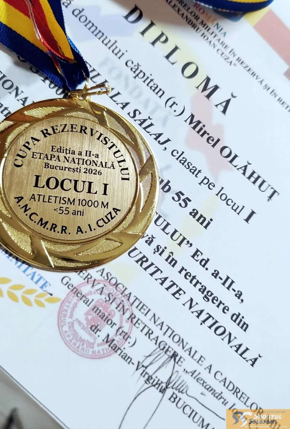 O medalie aurie cu inscripții este așezată pe o diplomă albă cu text imprimat negru și un sigiliu roz.
