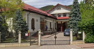 O clădire cu acoperiș roșu este înconjurată de copaci și un gard metalic negru în fața căreia se află o porțiță deschisă.