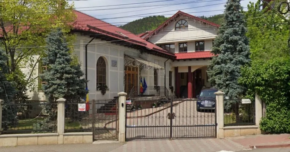 O clădire cu acoperiș roșu este înconjurată de copaci și un gard metalic negru în fața căreia se află o porțiță deschisă.