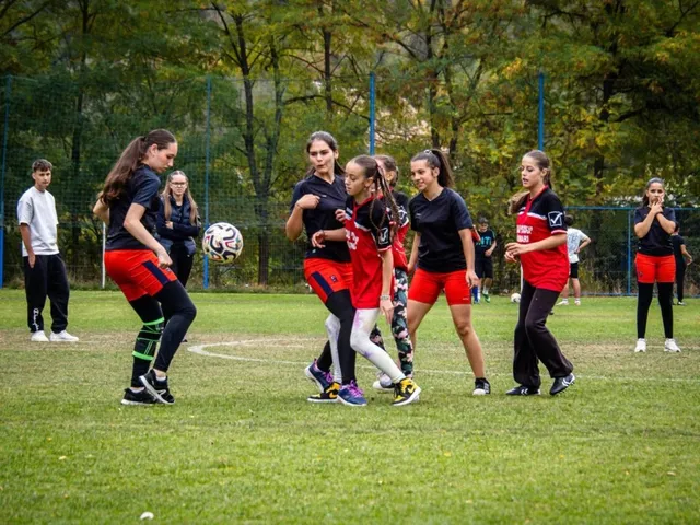 Fetele joacă fotbal pe un teren de iarbă verde, purtând echipamente sportive colorate, în timp ce un grup de persoane privește în fundal.