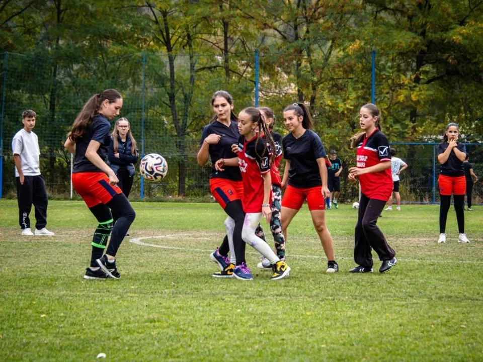 Fetele joacă fotbal pe un teren de iarbă verde, purtând echipamente sportive colorate, în timp ce un grup de persoane privește în fundal.