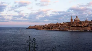 O vedere panoramică asupra unui oraș de coastă cu clădiri din piatră și o catedrală cu cupolă, situat lângă o mare întinsă sub un cer înnorat.