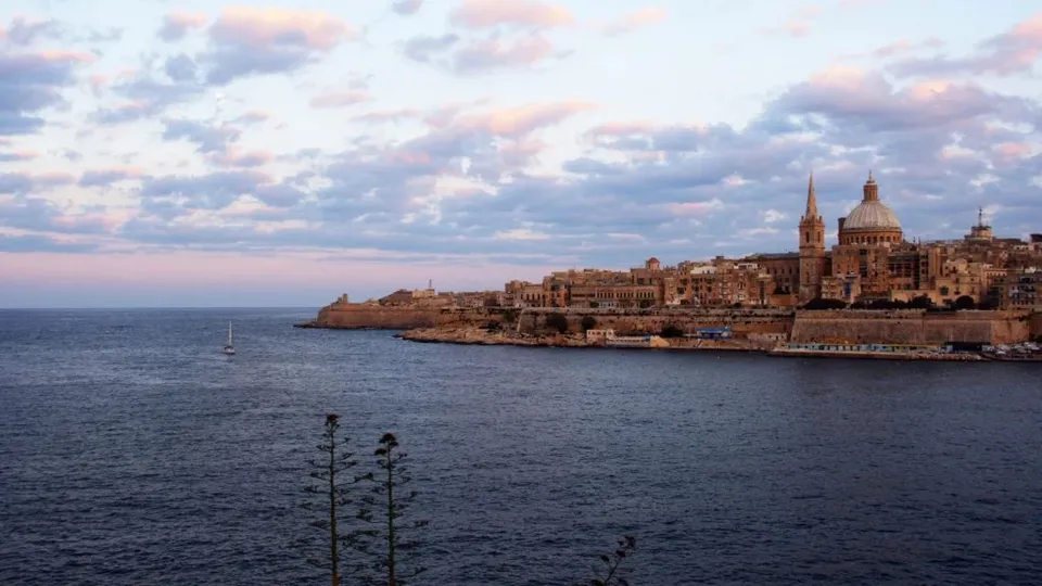 O vedere panoramică asupra unui oraș de coastă cu clădiri din piatră și o catedrală cu cupolă, situat lângă o mare întinsă sub un cer înnorat.